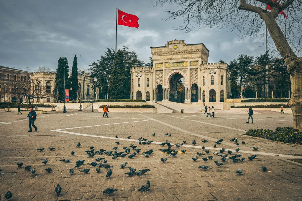 Istanbul University gate with pigeons and Turkish flag in a bustling city square.