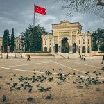 Istanbul University gate with pigeons and Turkish flag in a bustling city square.