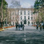 Students walking on a university campus surrounded by trees and spring flowers.