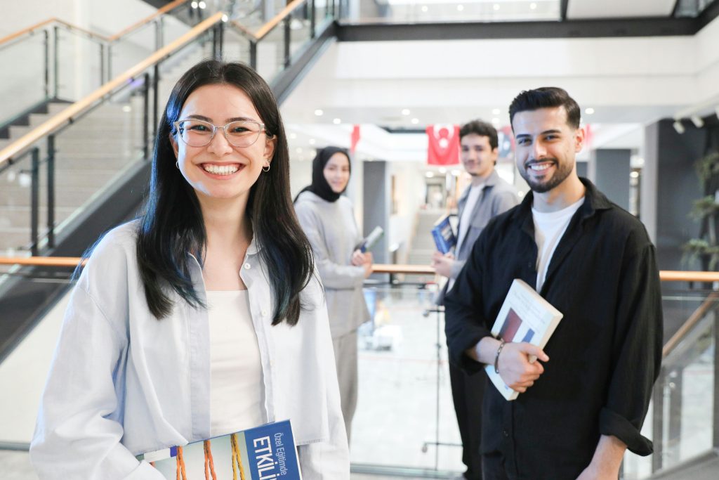 A diverse group of students in a modern indoor mall setting, smiling and holding books.