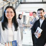A diverse group of students in a modern indoor mall setting, smiling and holding books.