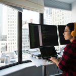 A businesswoman coding at a standing desk with city views through large windows, wearing headphones.