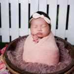 Adorable newborn with headband sleeping in pink wrap, captured in a cozy basket setting.