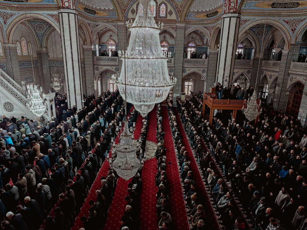 Aerial view of a large congregation praying inside an ornate mosque with chandeliers.