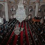Aerial view of a large congregation praying inside an ornate mosque with chandeliers.