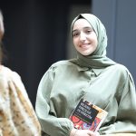 A young woman in a headscarf smiles while holding a book, indoors.