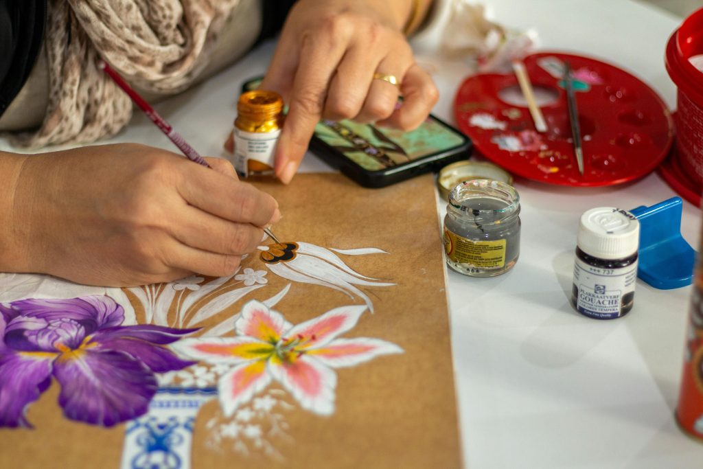 Artist painting vibrant flowers on paper using gouache paints in an indoor setting.