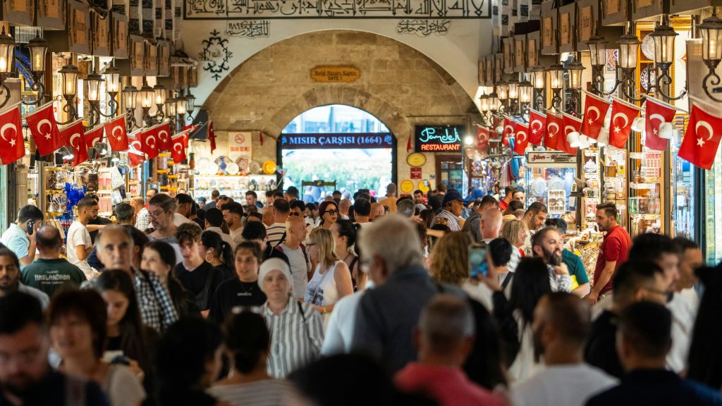 A crowded scene inside the vibrant Istanbul Spice Bazaar with people shopping and Turkish flags hanging.