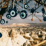 Nazar amulets on threads hanging from tree branches near rocky uneven formations with mountains and grass with plants in Turkey in Cappadocia region under gray cloudy sky in summer day
