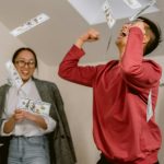 Two young adults in a room celebrating with falling dollar bills, expressing excitement and joy.