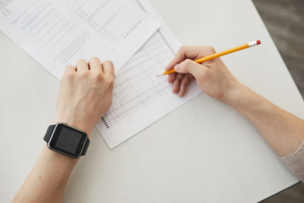 Unrecognizable student writing an exam using a pencil, watch visible on wrist.